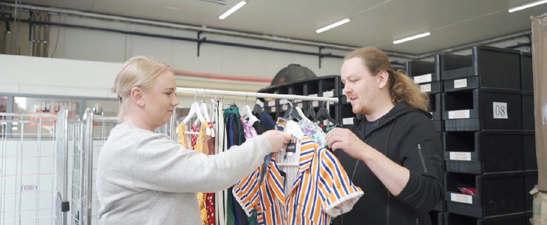 Kuvituskuva Miss Windy Shop -myymälästä. Taustalla vaaterekki. Omistajat pitävät vaatetta käsissään.