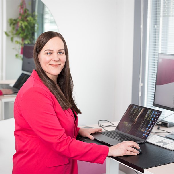 Woman working on a laptop at a standing desk, smiling at the camera