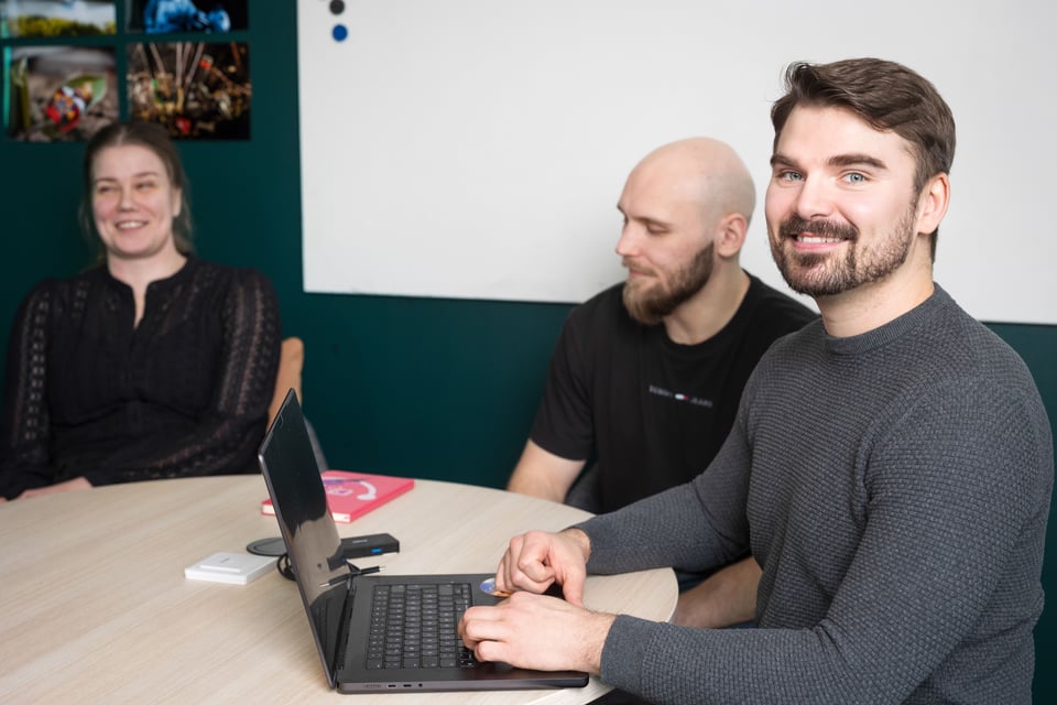 Three people sitting at a table, one using a laptop and smiling at the camera
